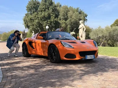 Corsista in ginocchio fotografa la Lotus Elise arancione davanti alla statua
