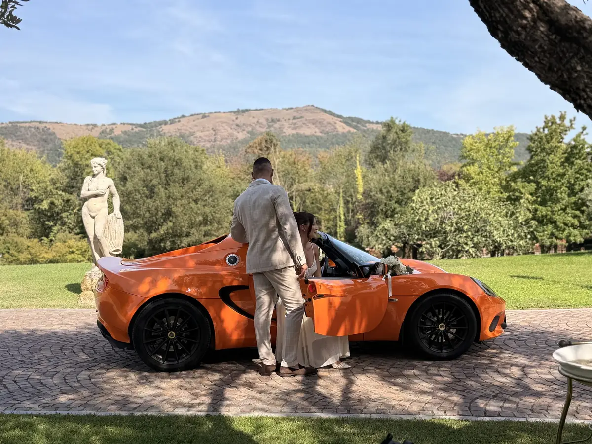 Sposi accanto alla Lotus Elise arancione tra le statue, con le montagne sullo sfondo